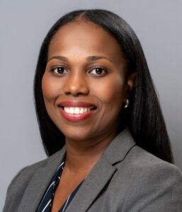 Woman in a gray blazer and striped shirt smiling at the camera against a plain gray background.