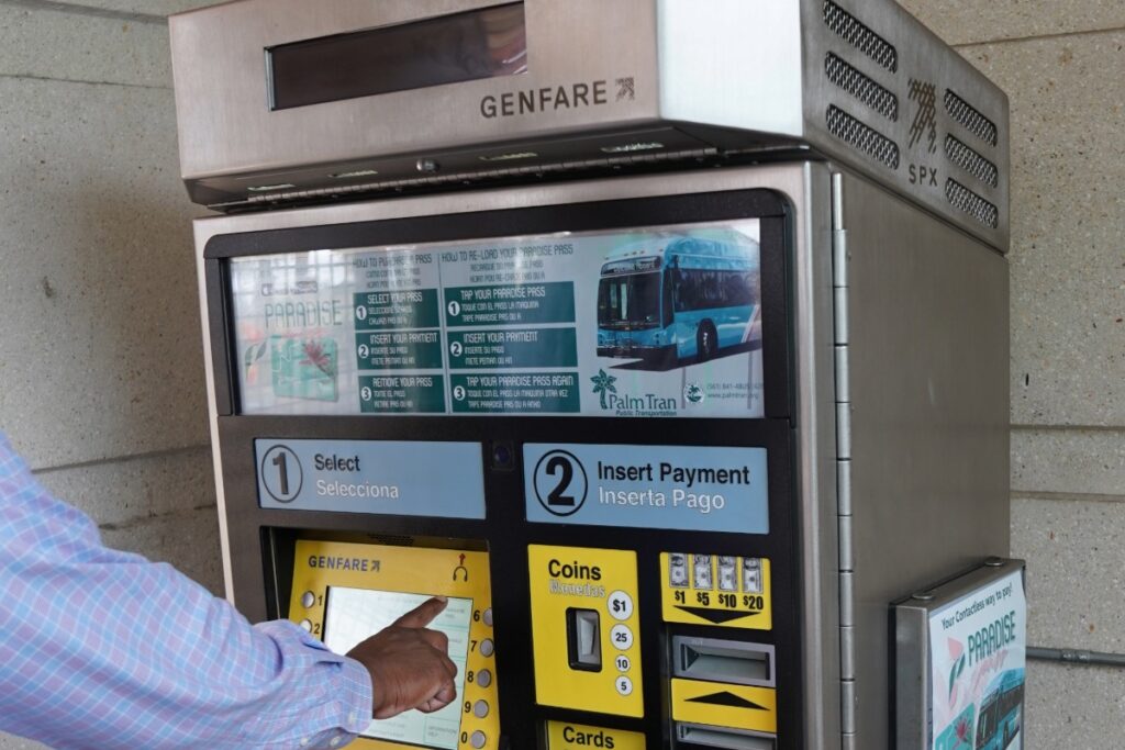 A person uses a Genfare ticket vending machine, pressing a button on the touchscreen to select and pay for public transit fare.