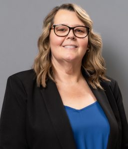 A woman with wavy, shoulder-length hair and glasses wears a black blazer over a blue top, standing against a plain gray background.